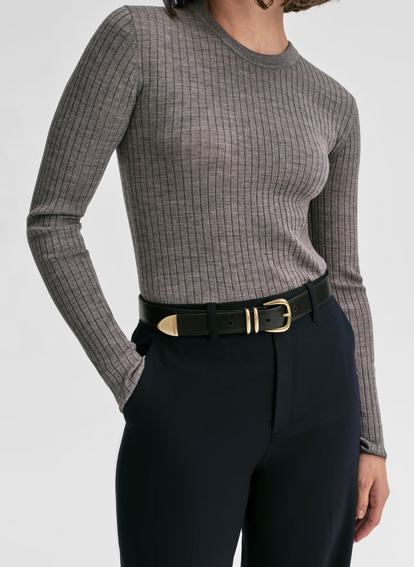 Person wearing a gray ribbed long-sleeve top and black pants with a gold belt on a light gray background