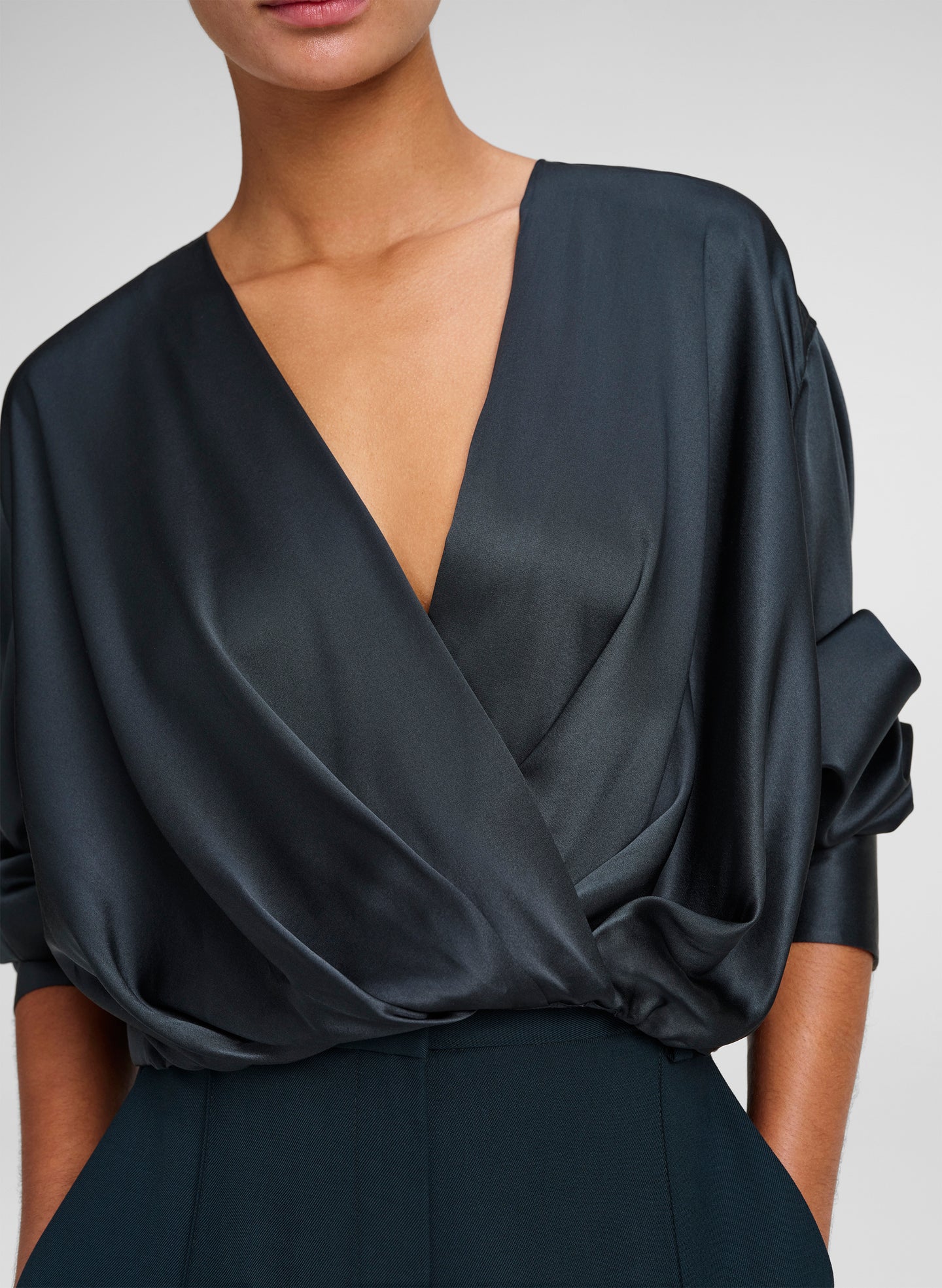 A close-up of a model wearing a dark green wrap-style blouse with a deep V-neckline and draped, voluminous sleeves.