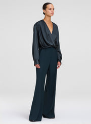 A model wearing a dark green wrap-style top with long sleeves and wide-leg pants, photographed against a neutral light gray studio background.