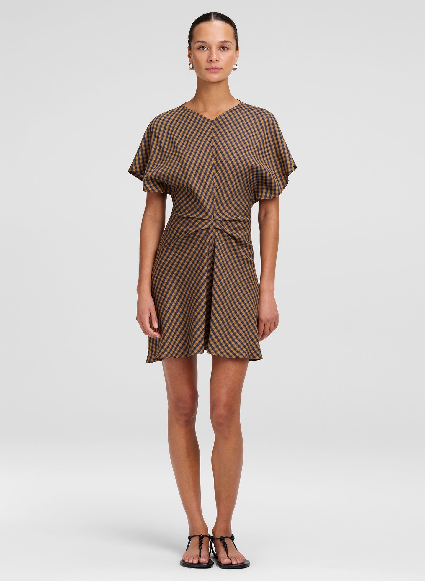A model wearing a brown and beige checkered mini dress with short flutter sleeves and a tie waist, standing against a light gray studio background.
