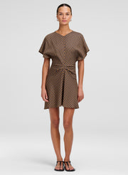 A model wearing a brown and beige checkered mini dress with short flutter sleeves and a tie waist, standing against a light gray studio background.