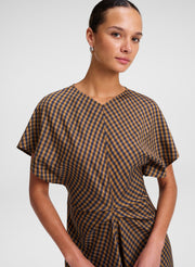 A woman wearing a brown and beige gingham dress with a V-neckline and flutter sleeves, photographed against a light gray studio background.