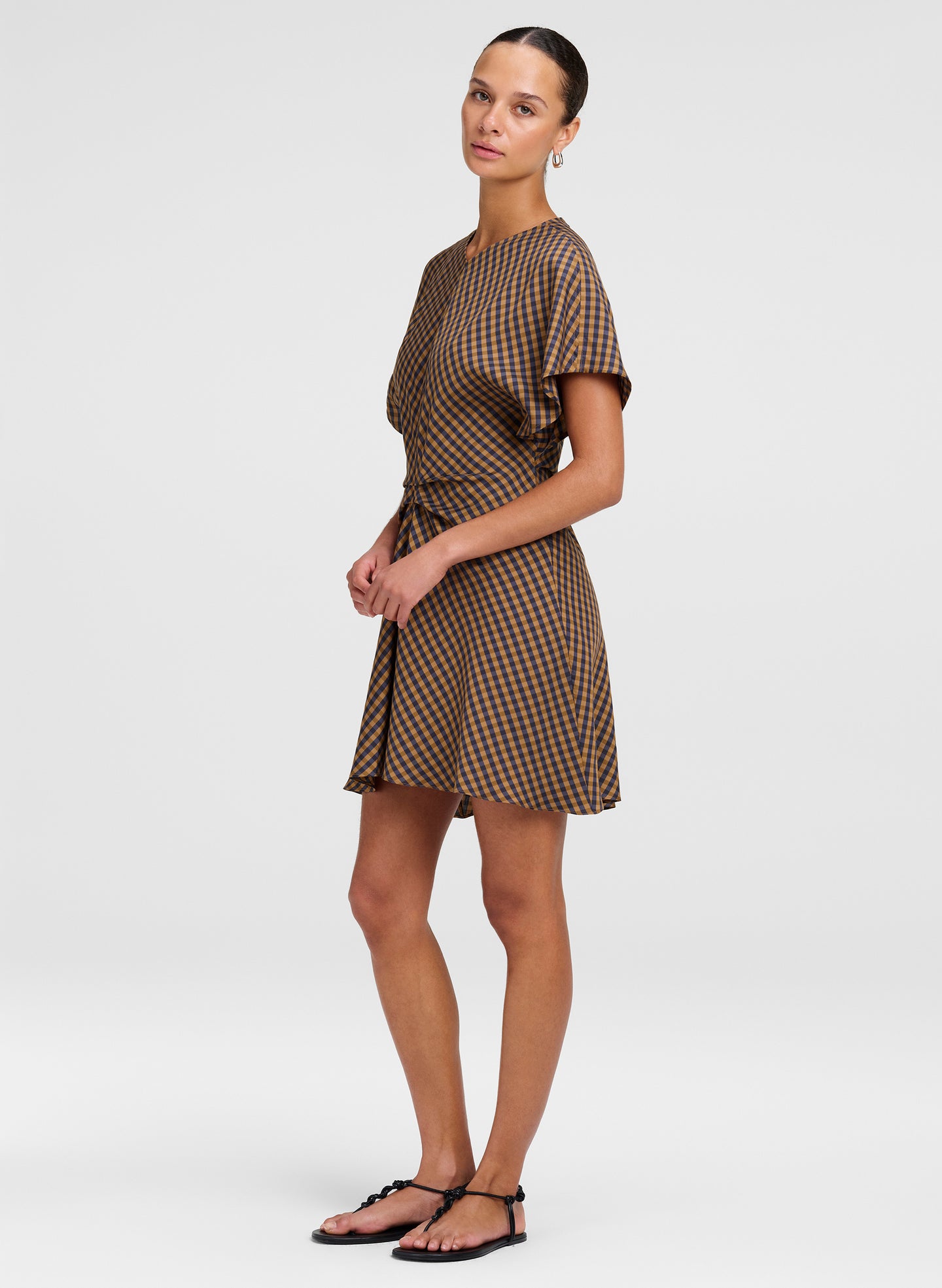 A woman modeling a brown and beige checkered mini dress with short sleeves and a tie waist, paired with black flat sandals, against a light gray studio background.