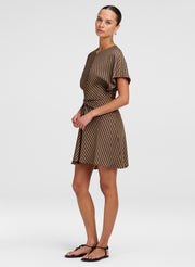 A woman modeling a brown and beige checkered mini dress with short sleeves and a tie waist, paired with black flat sandals, against a light gray studio background.