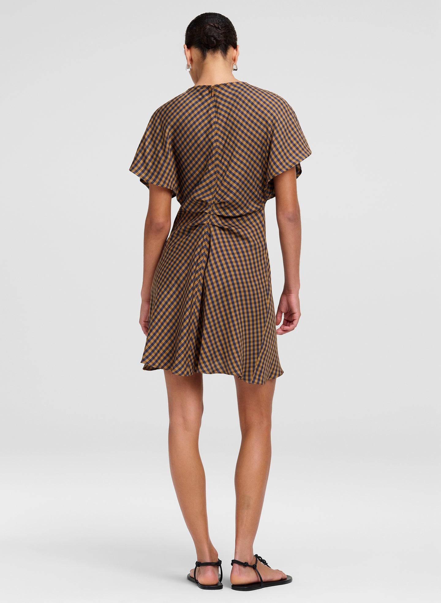 A woman viewed from the back wearing a brown and beige checkered mini dress with short flutter sleeves and a tie waist, paired with black flat sandals, against a light gray studio background.