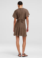 A woman viewed from the back wearing a brown and beige checkered mini dress with short flutter sleeves and a tie waist, paired with black flat sandals, against a light gray studio background.