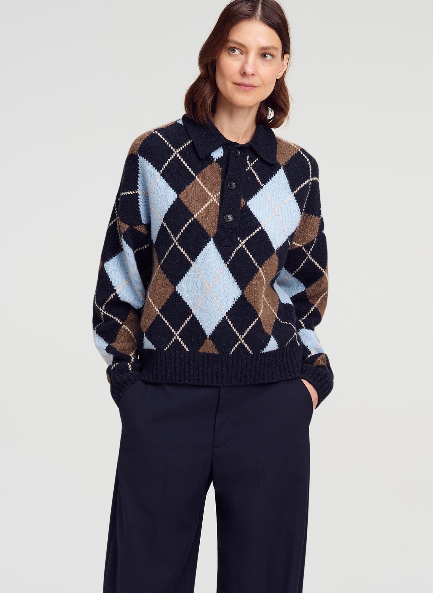 A woman wearing a navy, brown, and light blue argyle knit polo sweater with dark navy trousers, photographed against a light gray studio background.
