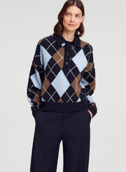 A woman wearing a navy, brown, and light blue argyle knit polo sweater with dark navy trousers, photographed against a light gray studio background.