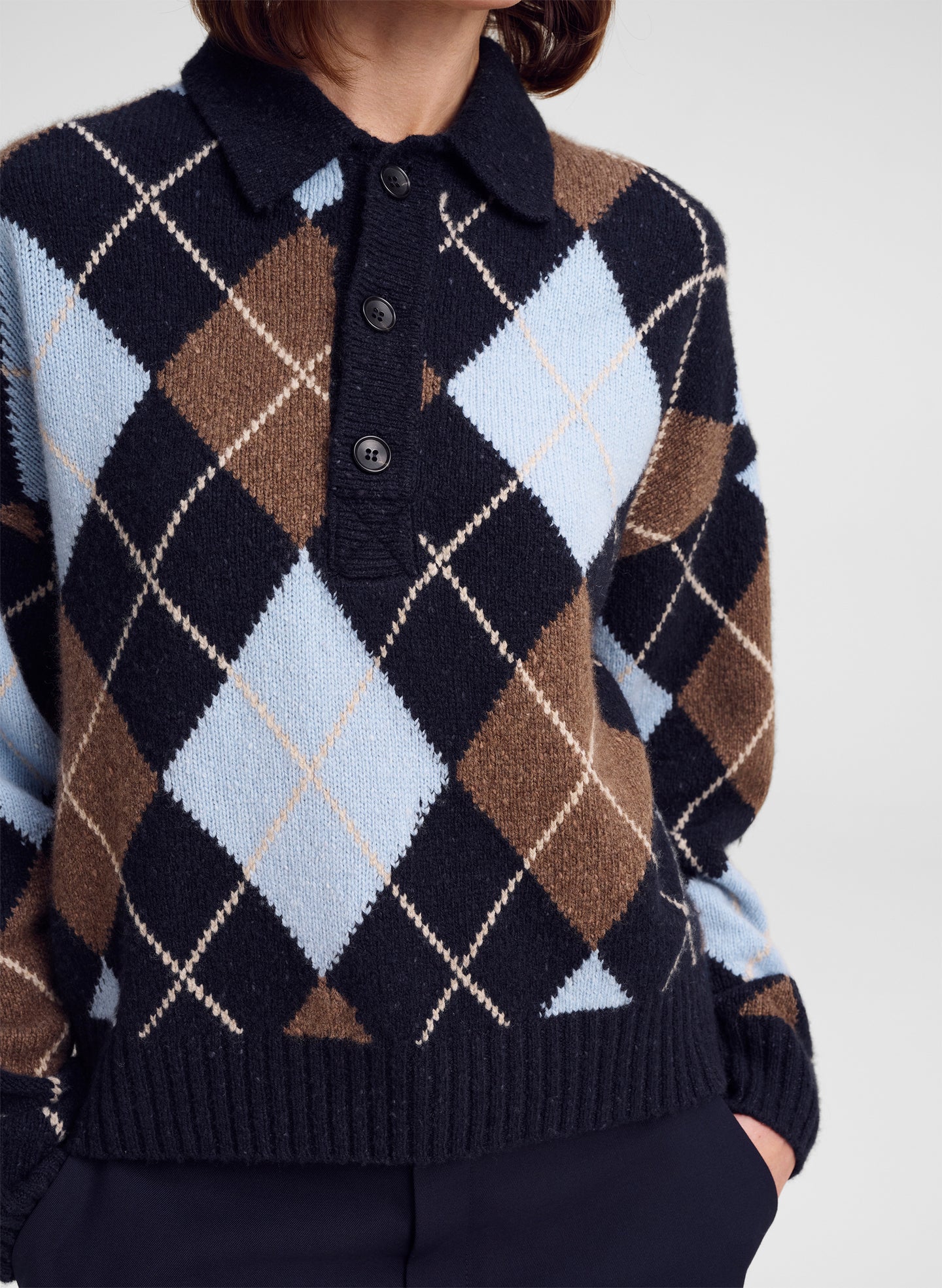 A close-up of a person wearing a knit polo sweater with an argyle pattern in navy, brown, light blue, and cream, paired with dark trousers against a light gray background.