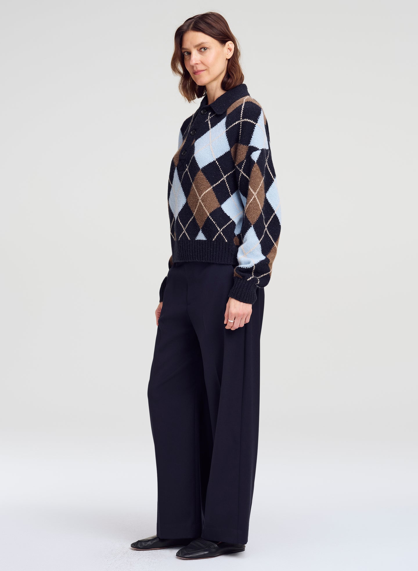 A woman modeling a navy blue argyle sweater with brown, beige, and light blue diamonds, paired with dark wide-leg trousers, against a light gray studio background.
