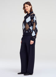 A woman modeling a navy blue argyle sweater with brown, beige, and light blue diamonds, paired with dark wide-leg trousers, against a light gray studio background.
