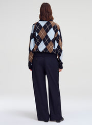 A model shown from the back wearing a cropped argyle sweater with navy, brown, beige, and white diamonds, paired with dark navy wide-leg trousers, against a light gray studio background.