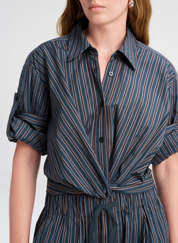 A close-up of a model wearing a dark blue and black vertically striped shirt  with a tie waist and rolled-up sleeves.