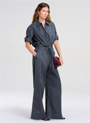 A woman models a dark blue and gray pinstriped shirt with wide-leg pants, holding a small burgundy clutch against a light gray studio background.