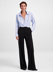 A model stands against a light gray studio background wearing a light blue long-sleeve button-up shirt tied at the waist and black wide-leg trousers.