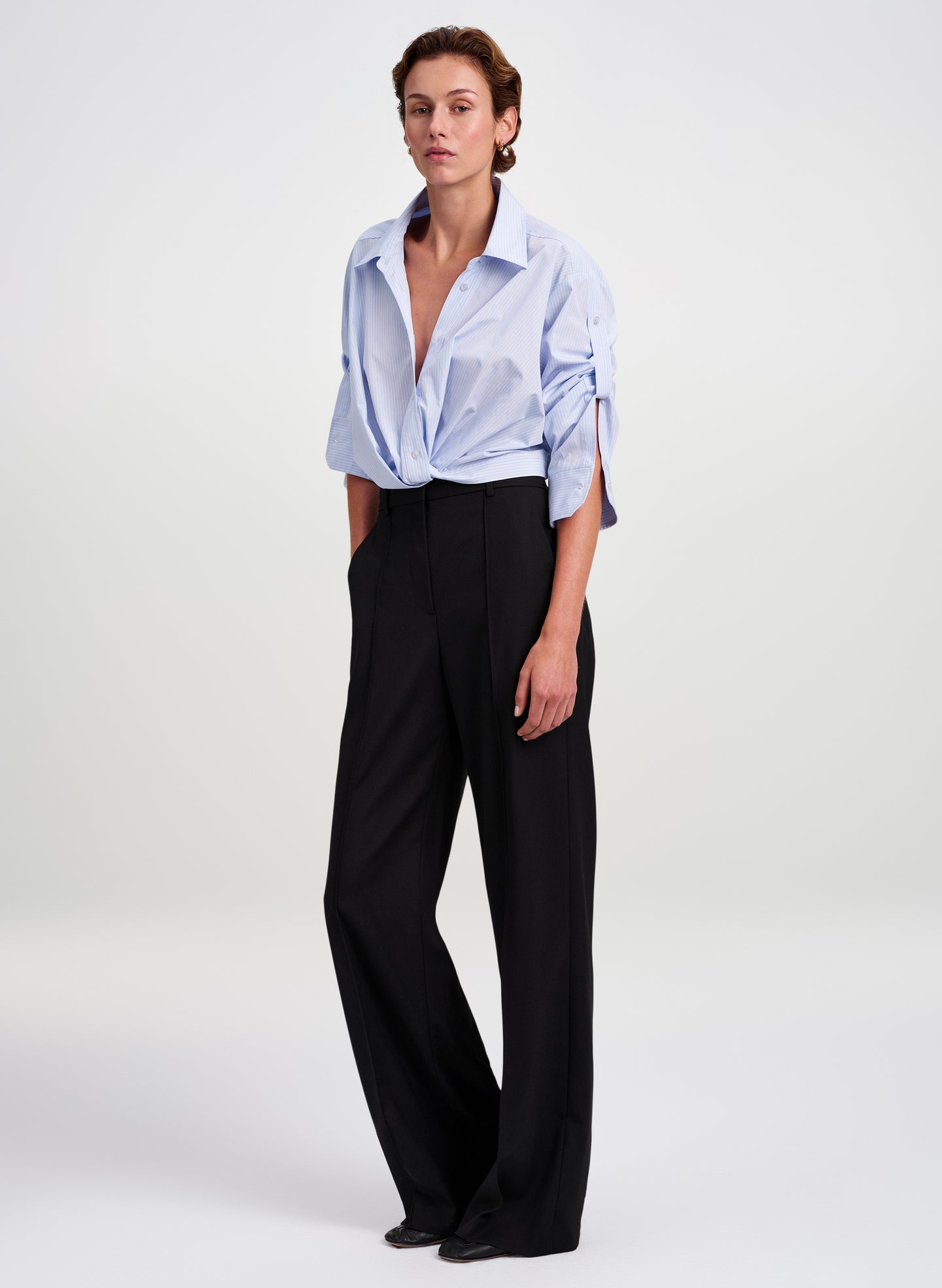 A model stands against a light gray studio background wearing a light blue button-down shirt tied at the waist and black wide-leg trousers.