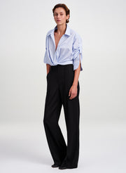 A model stands against a light gray studio background wearing a light blue button-down shirt tied at the waist and black wide-leg trousers.