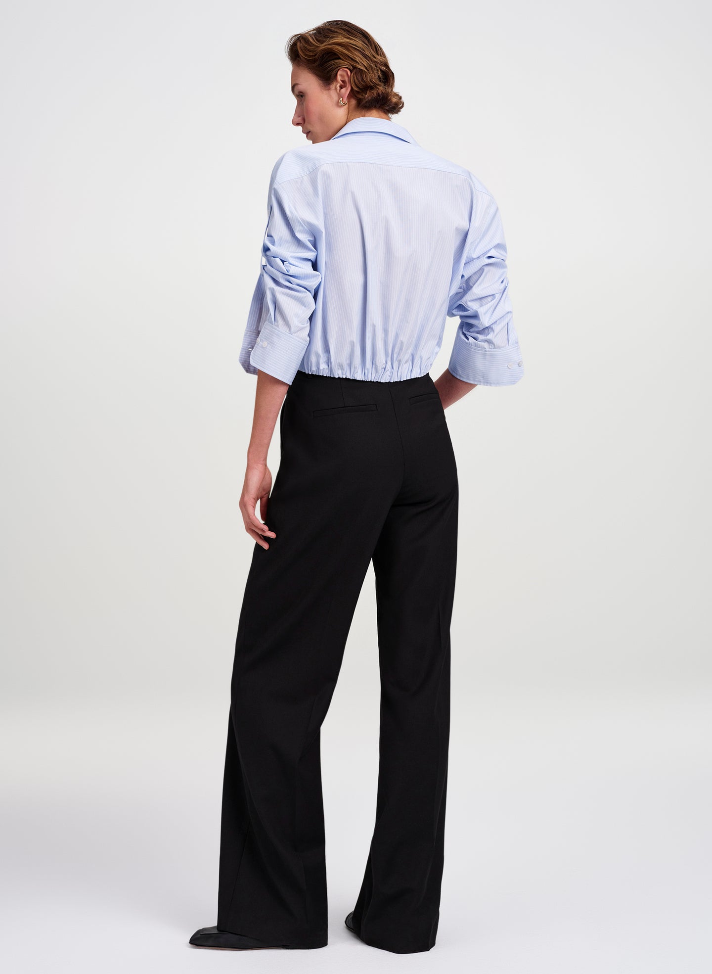 A model shown from the back wearing a light blue long-sleeve button-up shirt and black wide-leg trousers, photographed against a clean white studio background.