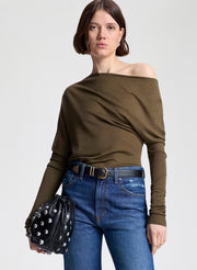 Woman wearing an off-shoulder olive green top and blue jeans, holding a black studded bag on a white background