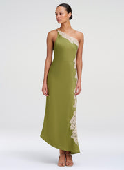 A woman modeling a one-shoulder olive green midi dress with cream lace trim along the neckline and side, photographed against a plain light gray studio background.