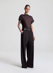 Woman wearing a brown blouse with black lace details and brown pants on a light gray background