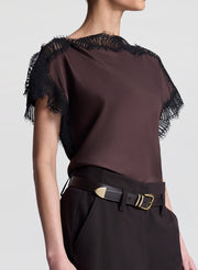 Woman wearing a brown blouse with black lace details and brown pants on a light gray background