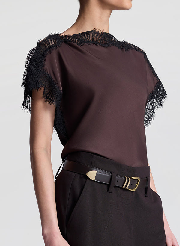 Woman wearing a brown blouse with black lace details and brown pants on a light gray background