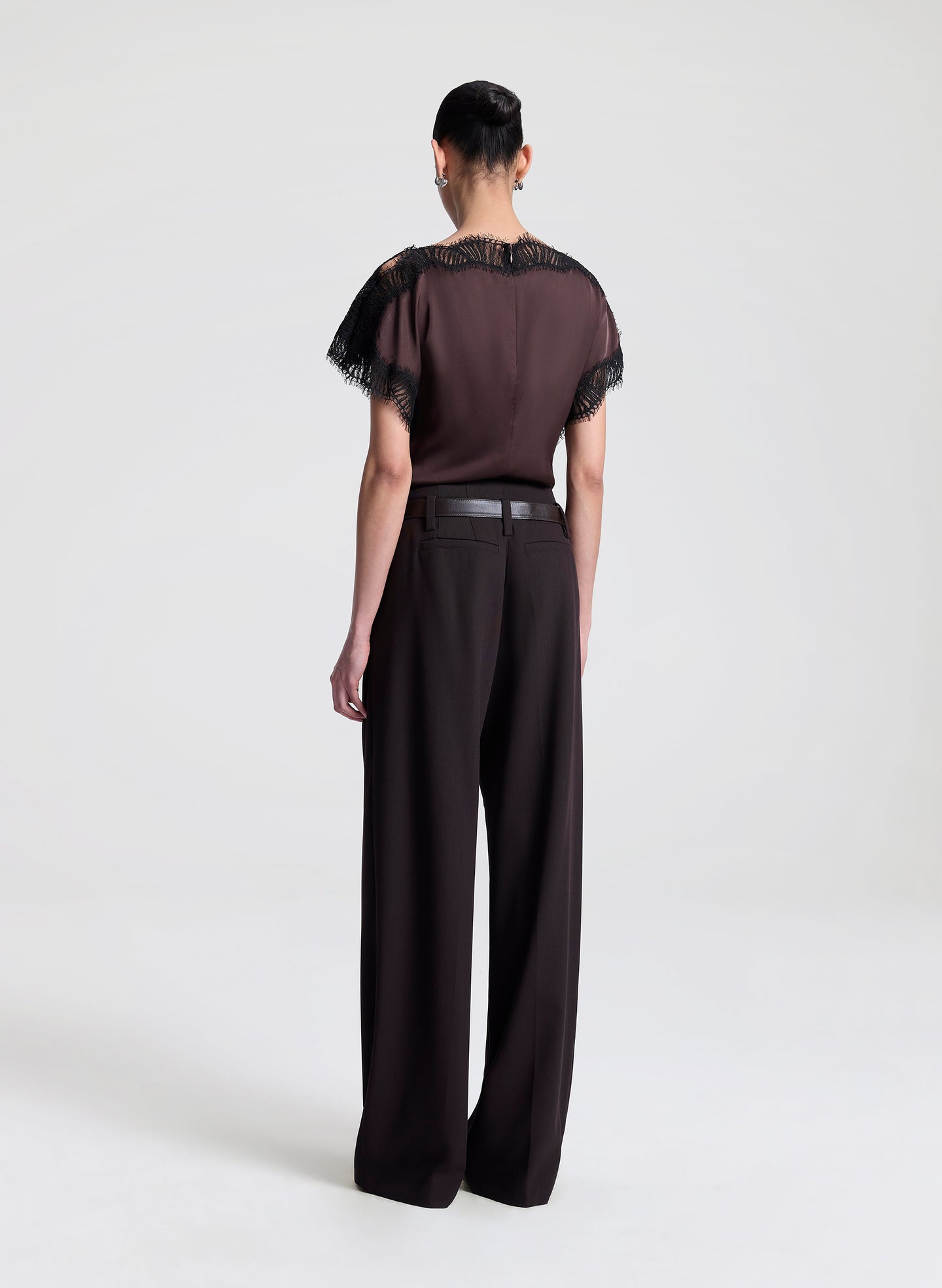 Woman wearing a brown blouse with black lace details and brown pants on a light gray background