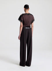 Woman wearing a brown blouse with black lace details and brown pants on a light gray background