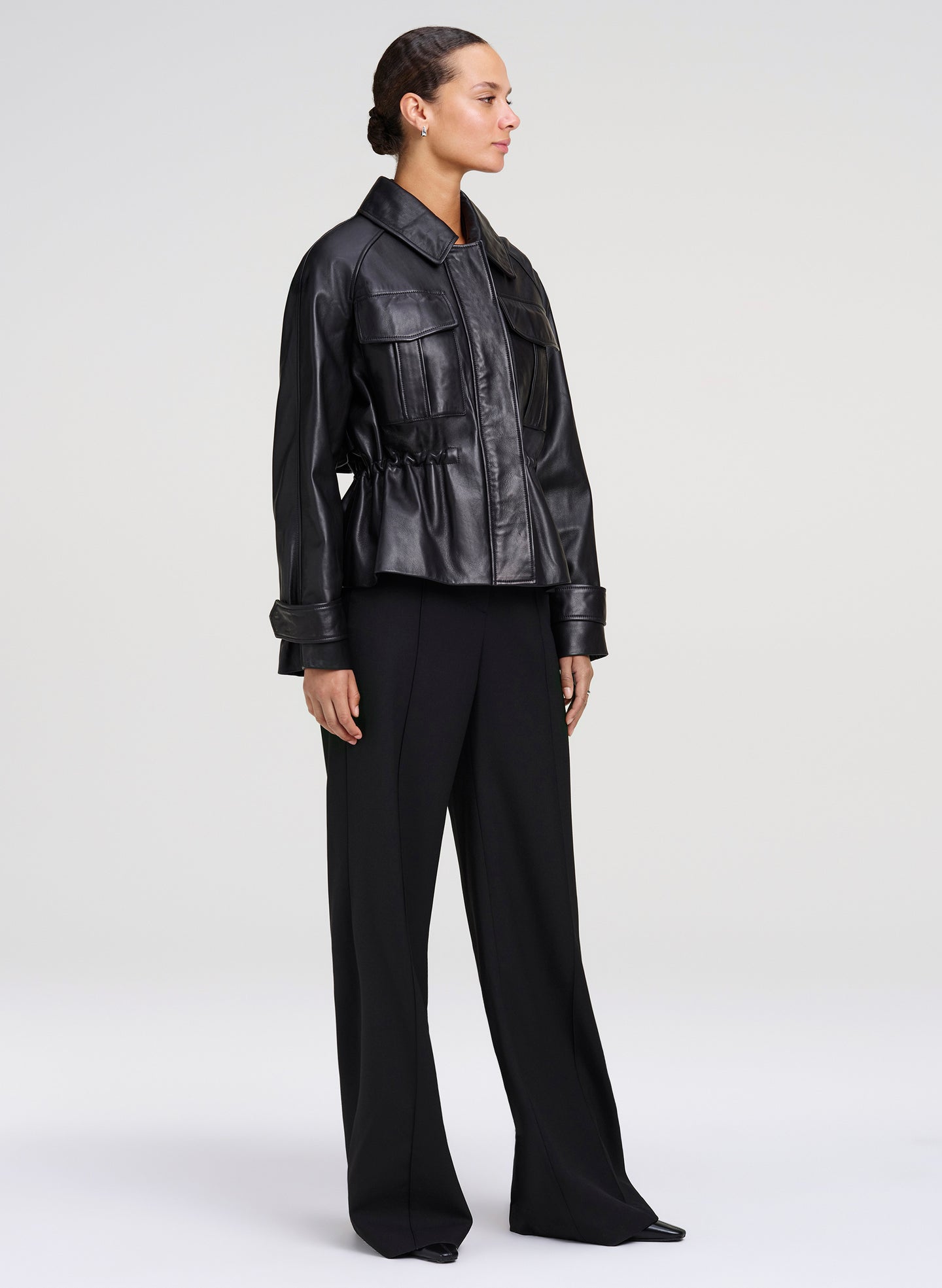 A model is shown from the side wearing a black leather jacket with a peplum waist and wide-leg black trousers, photographed against a neutral light gray studio background.