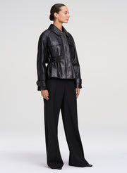 A model is shown from the side wearing a black leather jacket with a peplum waist and wide-leg black trousers, photographed against a neutral light gray studio background.