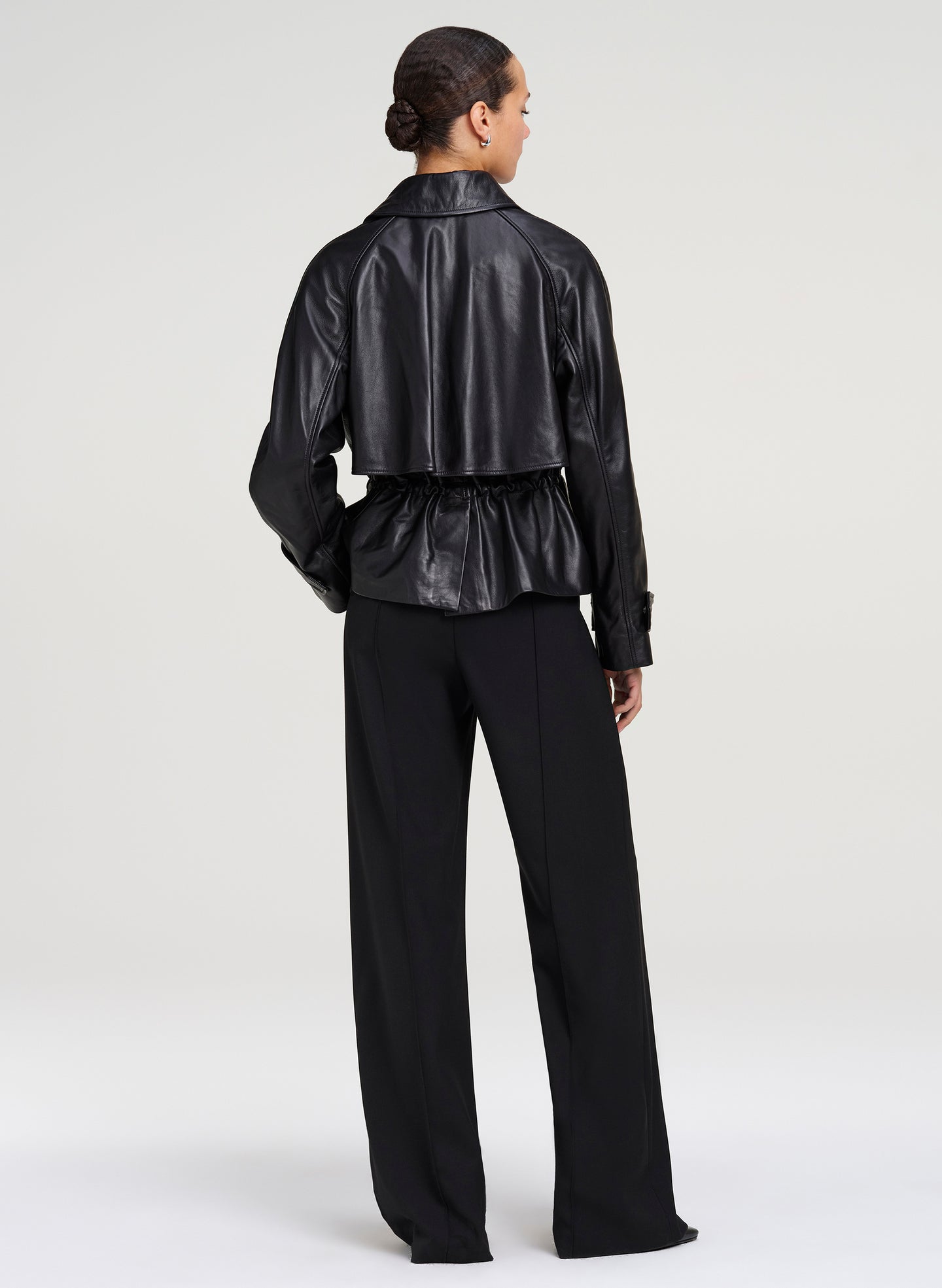 A model shown from the back wearing a black cropped leather jacket with a peplum waist and black wide-leg trousers, set against a plain light gray studio background.