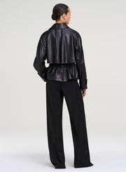 A model shown from the back wearing a black cropped leather jacket with a peplum waist and black wide-leg trousers, set against a plain light gray studio background.