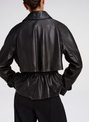 Rear view of a model wearing a black leather cropped jacket with a gathered waist and collar, paired with black trousers, against a light gray studio background.