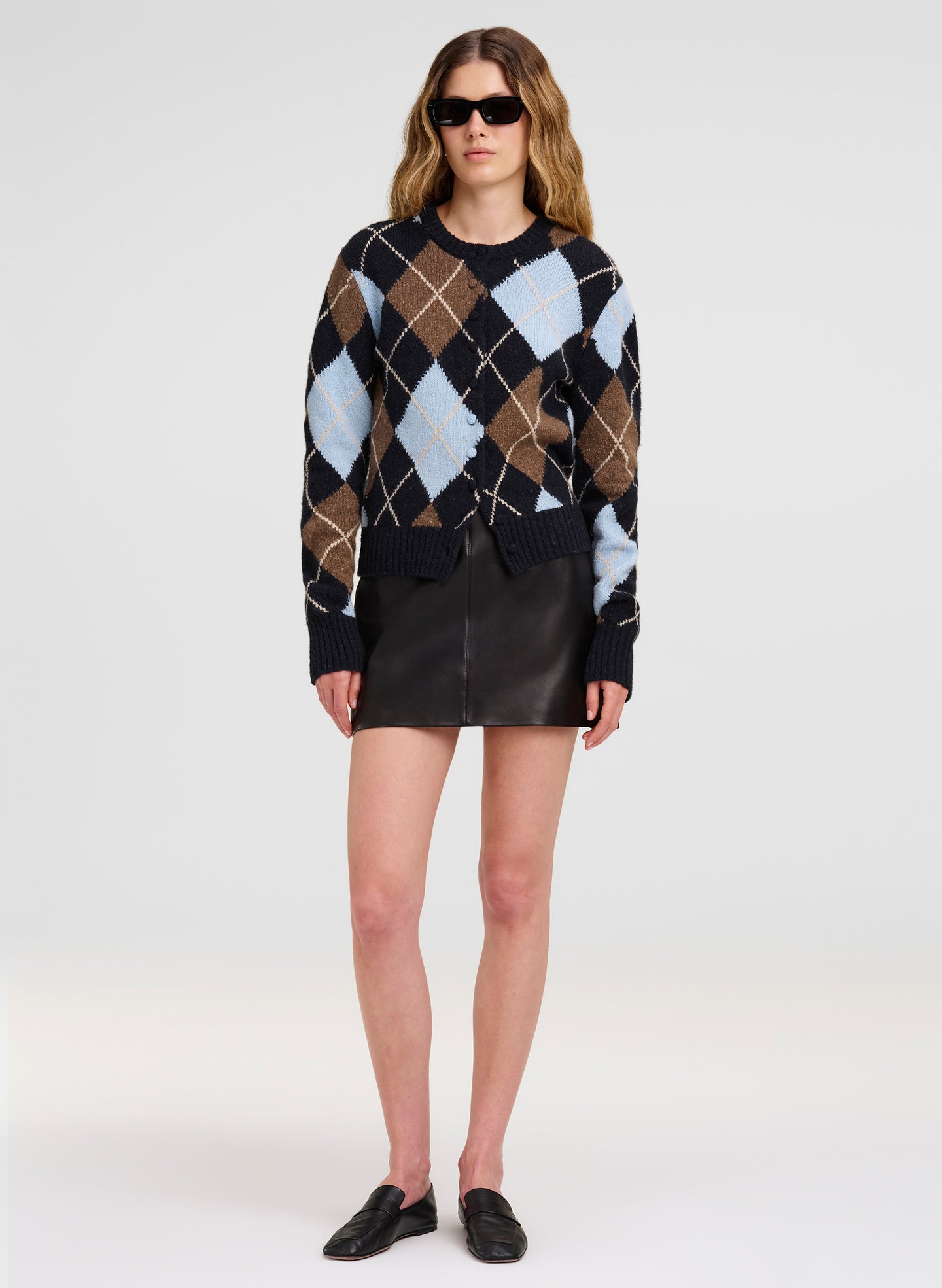 A model wearing a black cardigan sweater with a brown, beige, and light blue argyle pattern, paired with a black leather mini skirt and black loafers, on a light gray studio background.