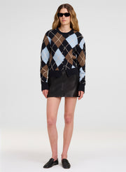 A model wearing a black cardigan sweater with a brown, beige, and light blue argyle pattern, paired with a black leather mini skirt and black loafers, on a light gray studio background.