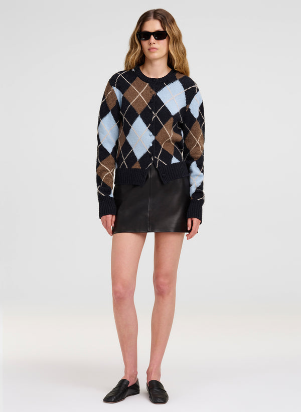 A model wearing a black cardigan sweater with a brown, beige, and light blue argyle pattern, paired with a black leather mini skirt and black loafers, on a light gray studio background.
