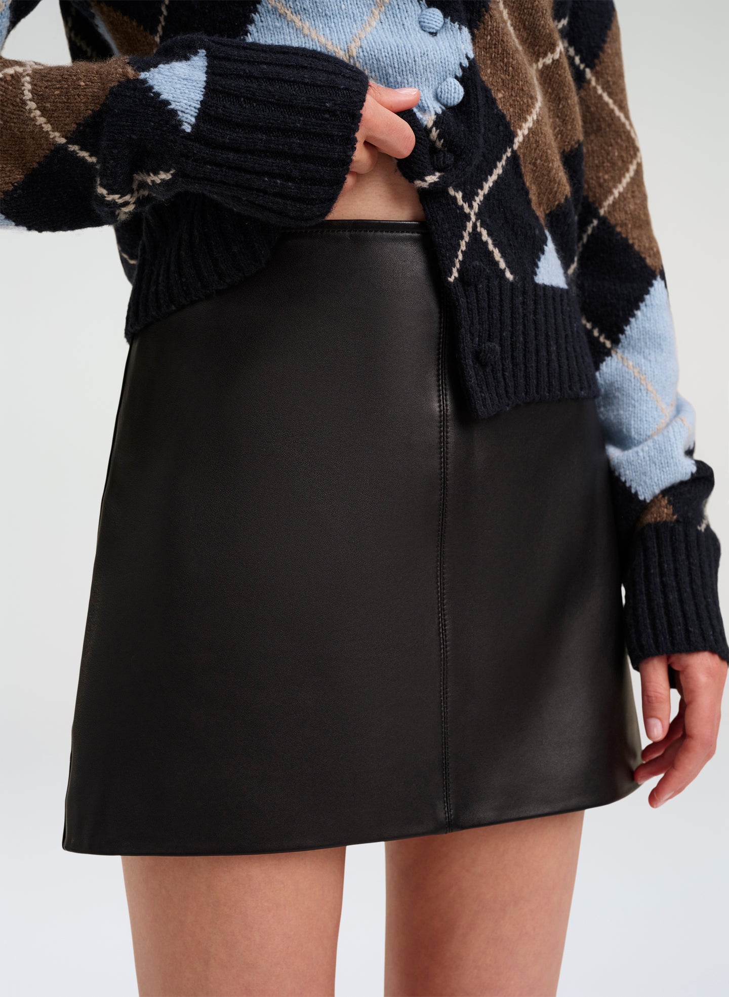 A model wearing a black leather A-line mini skirt paired with a brown, black, and light blue argyle knit cardigan.