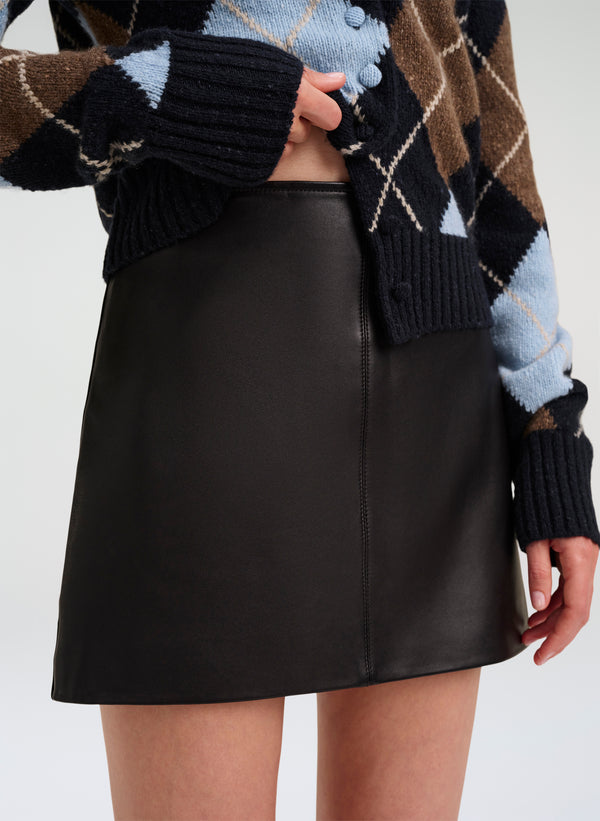 A model wearing a black leather A-line mini skirt paired with a brown, black, and light blue argyle knit cardigan.