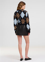 A woman shown from the back wearing a black, brown, and light blue argyle sweater with a black mini skirt and black flats, photographed against a light gray studio background.