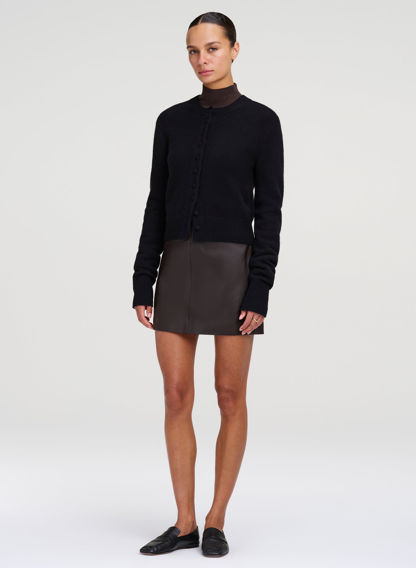 A model stands against a light gray studio background wearing a black long-sleeve mock-neck knit top, a dark brown mini skirt, and black slip-on shoes.