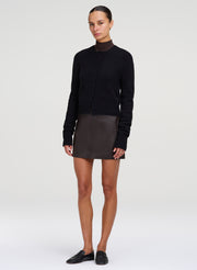 A model stands against a light gray studio background wearing a black long-sleeve mock-neck knit top, a dark brown mini skirt, and black slip-on shoes.