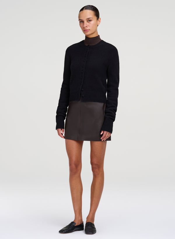 A model stands against a light gray studio background wearing a black long-sleeve mock-neck knit top, a dark brown mini skirt, and black slip-on shoes.