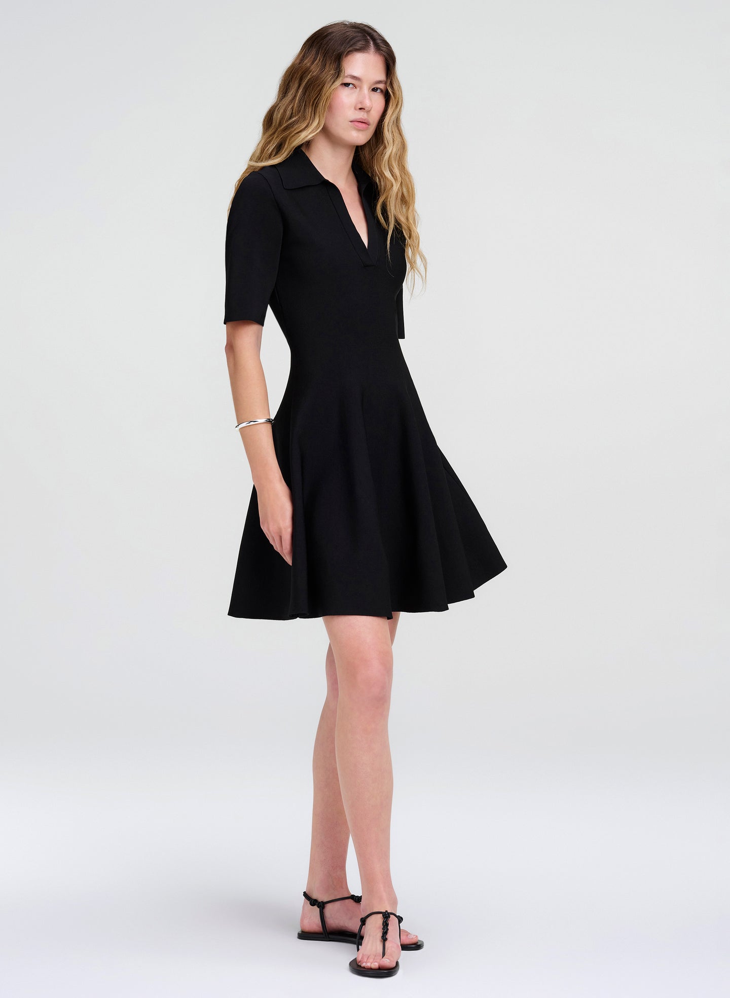 A woman modeling a black short-sleeve fit-and-flare mini dress with a V-neckline, paired with black strappy flat sandals, against a light gray studio background.