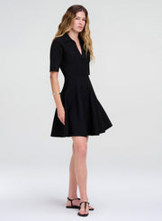 A woman modeling a black short-sleeve fit-and-flare mini dress with a V-neckline, paired with black strappy flat sandals, against a light gray studio background.