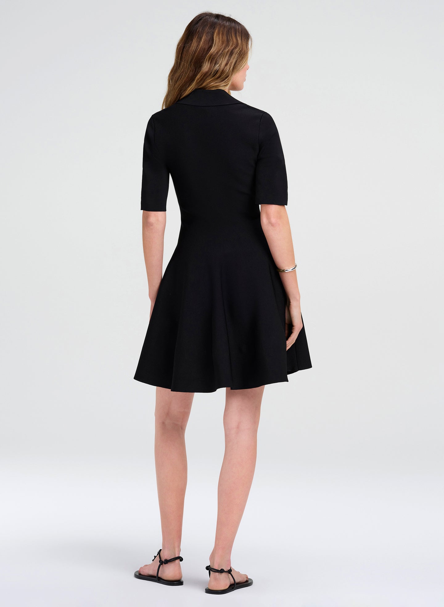 A woman viewed from the back wearing a black short-sleeve collared dress with a flared skirt, paired with black strappy sandals, against a light gray studio background.