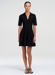 A woman modeling a black short-sleeve collared shift dress with a V-neckline and flared skirt, paired with black strappy sandals, against a light gray studio background.