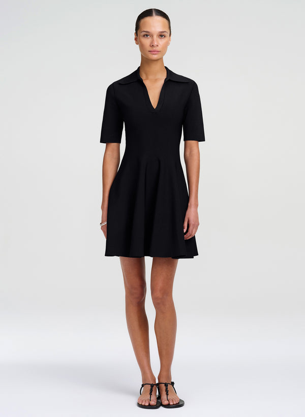 A woman modeling a black short-sleeve collared shift dress with a V-neckline and flared skirt, paired with black strappy sandals, against a light gray studio background.