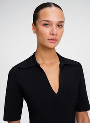 A close-up portrait of a model wearing a black short-sleeve collared knit top with a deep V-neckline, set against a plain light gray background.