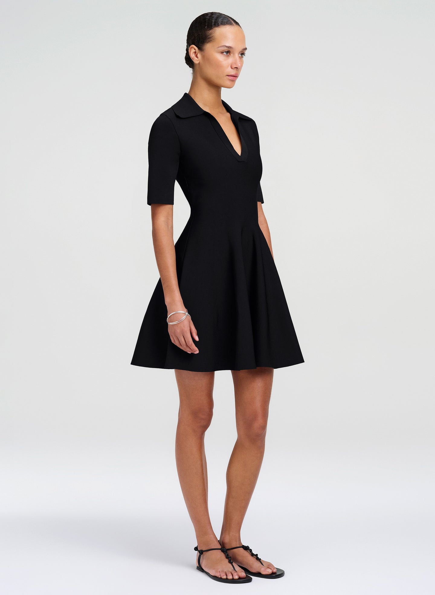 A woman modeling a black short-sleeve collared mini dress with a flared skirt, shown against a plain light gray studio background.